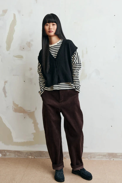A model stands against a textured, off-white plaster wall wearing a black knit vest over a black and white striped long-sleeve shirt, paired with dark brown corduroy pants and dark blue slip-on shoes.