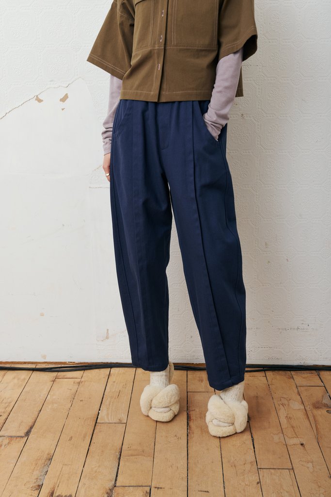 A person wearing loose navy blue relaxed-fit trousers, styled with a brown short-sleeve button-up shirt, a light purple long-sleeve layer, and cream fuzzy slippers, standing on a wooden floor against a white wall.