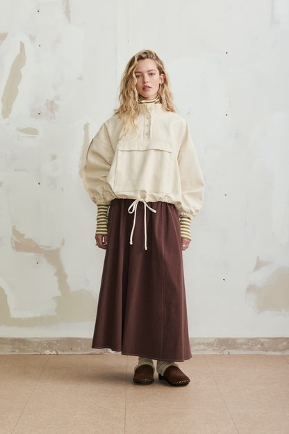 A model stands against a textured, off-white plaster wall wearing a cream-colored long-sleeve blouse with a ruffled collar and a dark brown midi skirt.