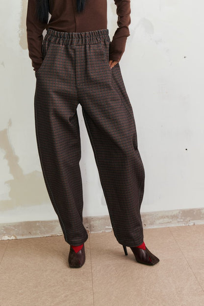 A person wearing loose, wide-leg brown plaid trousers with an elastic waistband, paired with a brown top and dark pointed-toe heels, standing against a light off-white wall.