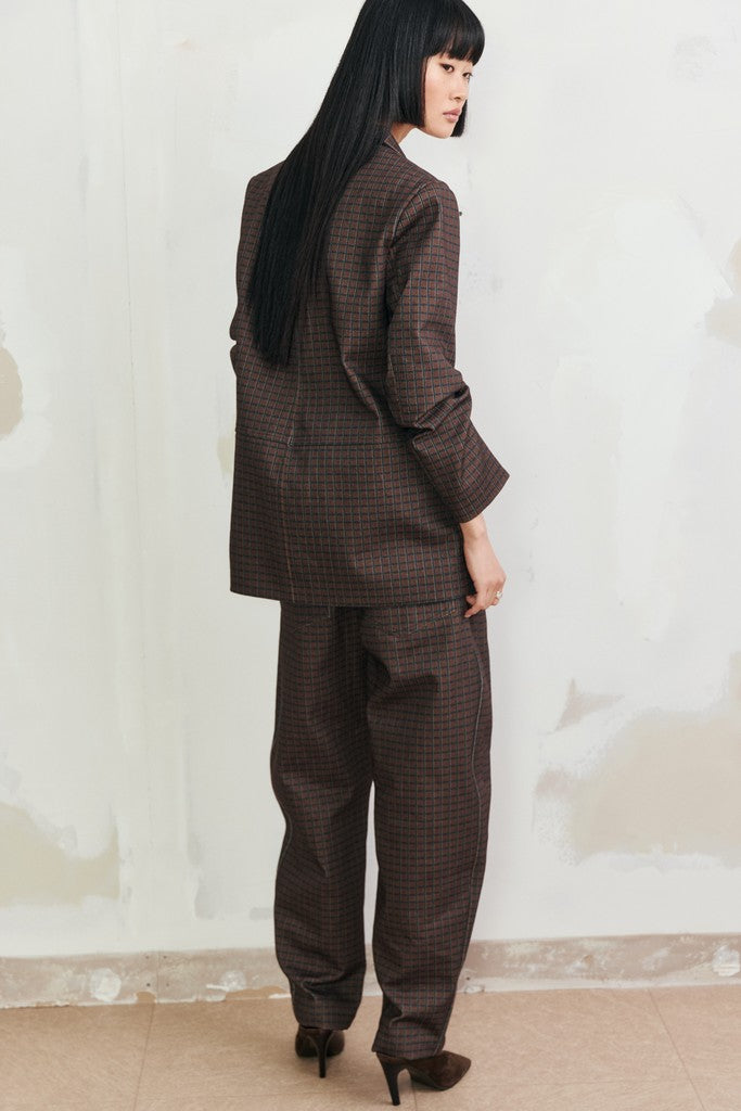 A model with long black hair is shown from the back wearing a dark brown and black checkered blazer and matching wide-leg trousers, standing against a textured off-white wall.