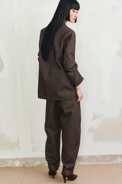 A model with long black hair is shown from the back wearing a dark brown and black checkered blazer and matching wide-leg trousers, standing against a textured off-white wall.