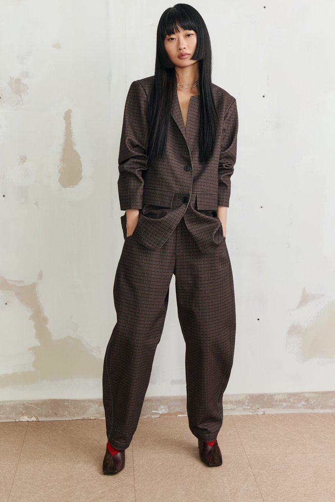 A model with long black hair stands against a textured off-white wall wearing a dark brown checkered two-piece suit consisting of a blazer and matching trousers, paired with dark brown shoes.