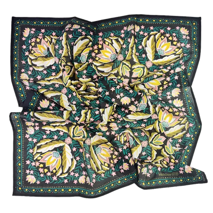 A square silk scarf featuring a botanical pattern with large yellow and green leaves, pink flowers, and a dark border, laid flat on a white background.