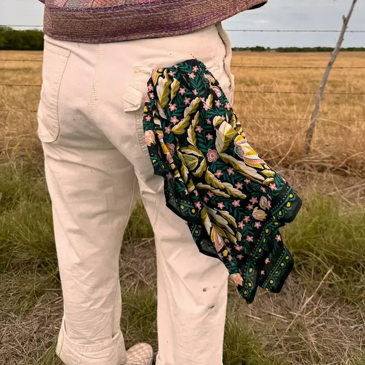 A person standing outdoors in a field, wearing cream pants and a patterned top, with a dark green embroidered scarf draped over the back of the pants. The scarf features yellow, pink, and green floral and leaf embroidery.