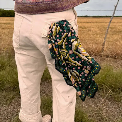A person standing outdoors in a field, wearing cream pants and a patterned top, with a dark green embroidered scarf draped over the back of the pants. The scarf features yellow, pink, and green floral and leaf embroidery.