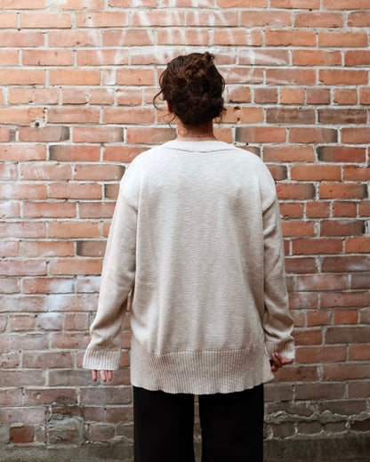 A person viewed from behind wearing a light beige knit sweater and black pants, standing in front of a red brick wall.