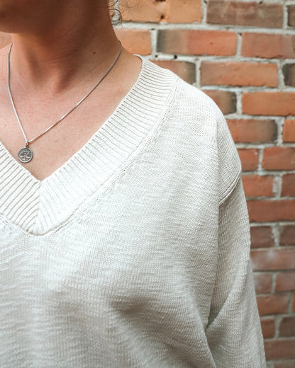 A close-up of a person wearing a light beige ribbed knit V-neck sweater and a silver pendant necklace, standing in front of a red brick wall.