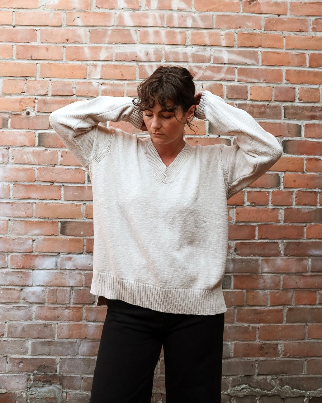 A woman wearing a cream-colored knit V-neck sweater and black pants stands against a red brick wall, adjusting her hair with both hands.