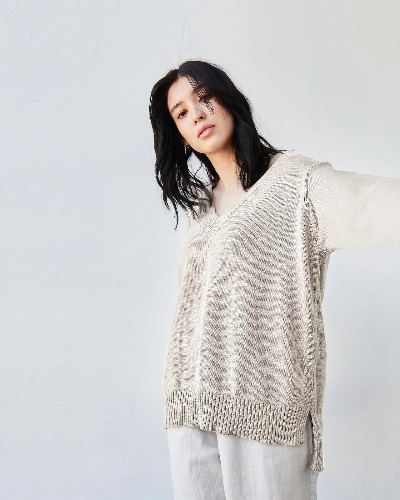 A woman wearing a loose, oversized beige knit sweater with a ribbed hem and light gray wide-leg pants, posed against a plain light gray wall.