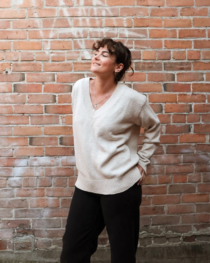 A woman wearing a cream-colored v-neck knit sweater and black pants stands against a red-brown brick wall.