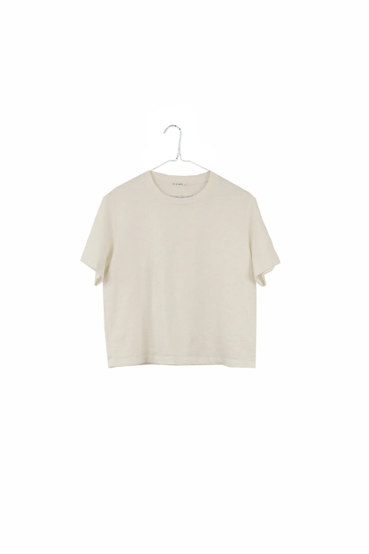A short-sleeve crewneck t-shirt in a light cream or off-white color, hanging on a simple metal hanger against a solid white background.