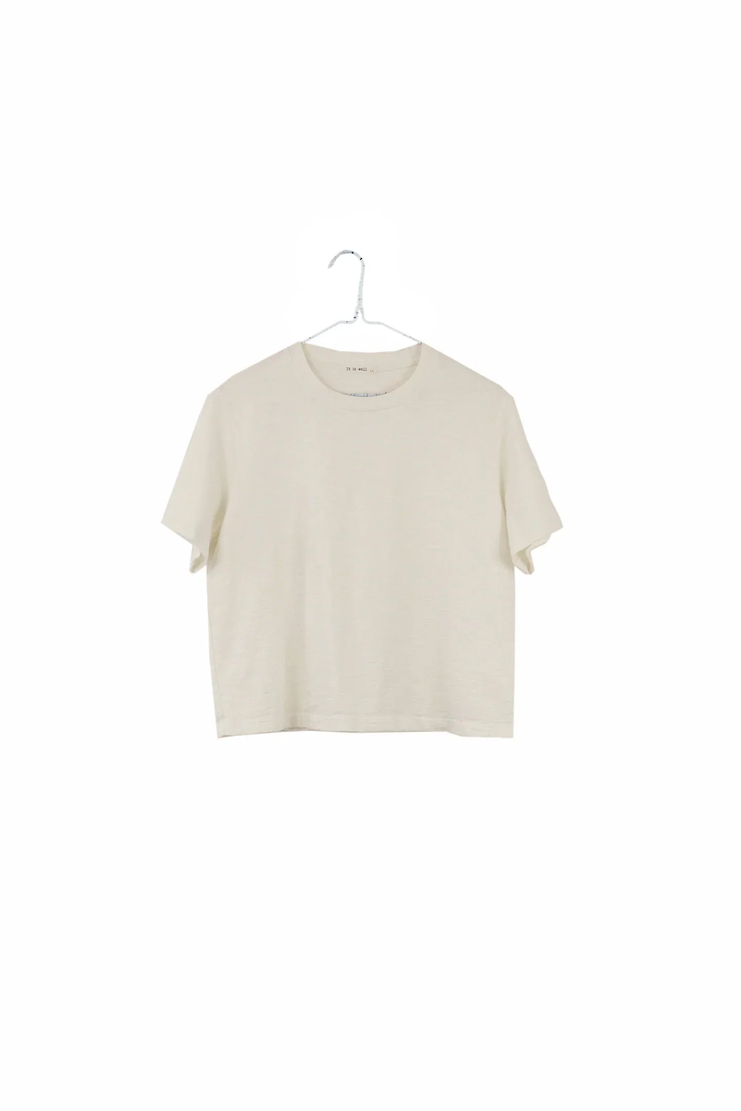 A short-sleeve crewneck t-shirt in a light cream or off-white color, hanging on a simple metal hanger against a solid white background.