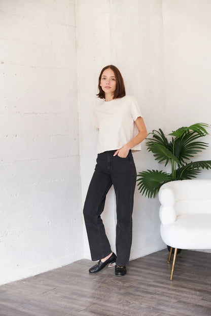 A woman stands in a bright, modern room wearing a cream short-sleeve top, black wide-leg pants, and black flats. She is positioned next to a white armchair and a green potted plant against a white textured wall.