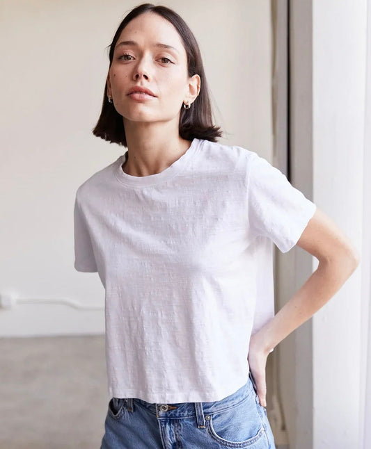 A woman with short dark hair wearing a light gray short-sleeve crewneck t-shirt and blue jeans, standing in a brightly lit indoor setting.