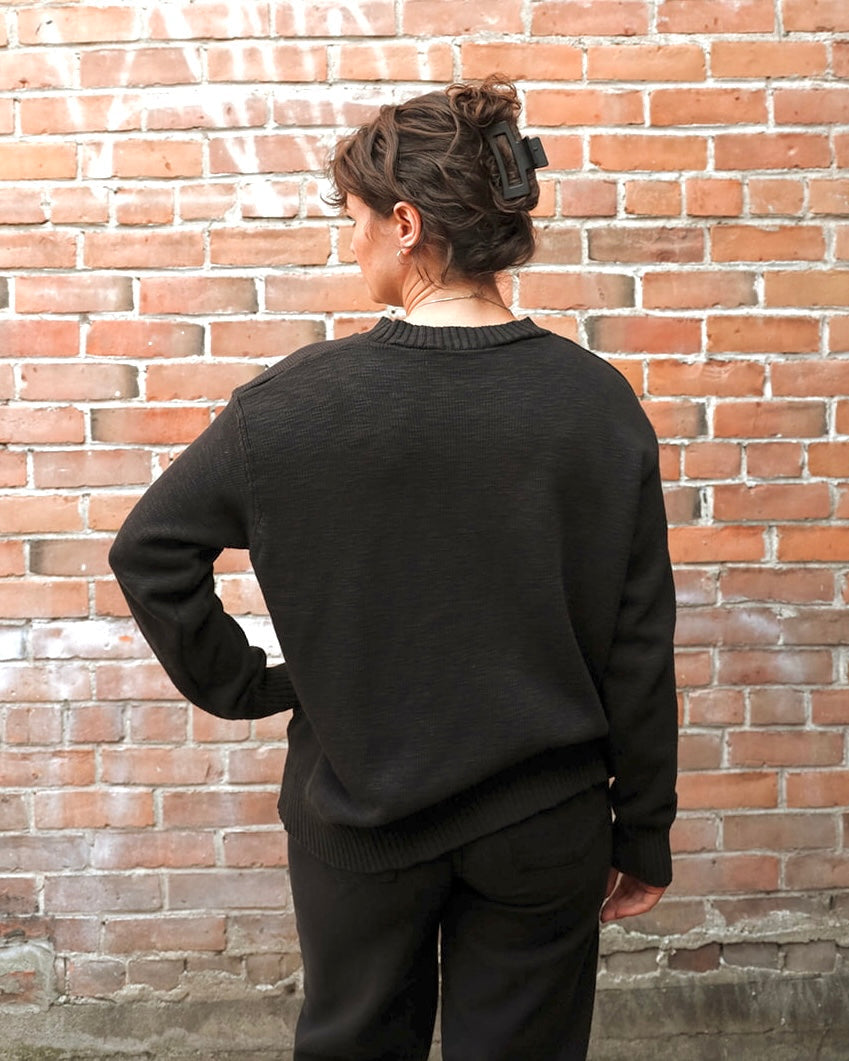 A person viewed from behind wearing a black knit crewneck sweater and matching black pants, standing in front of a red-brown brick wall.