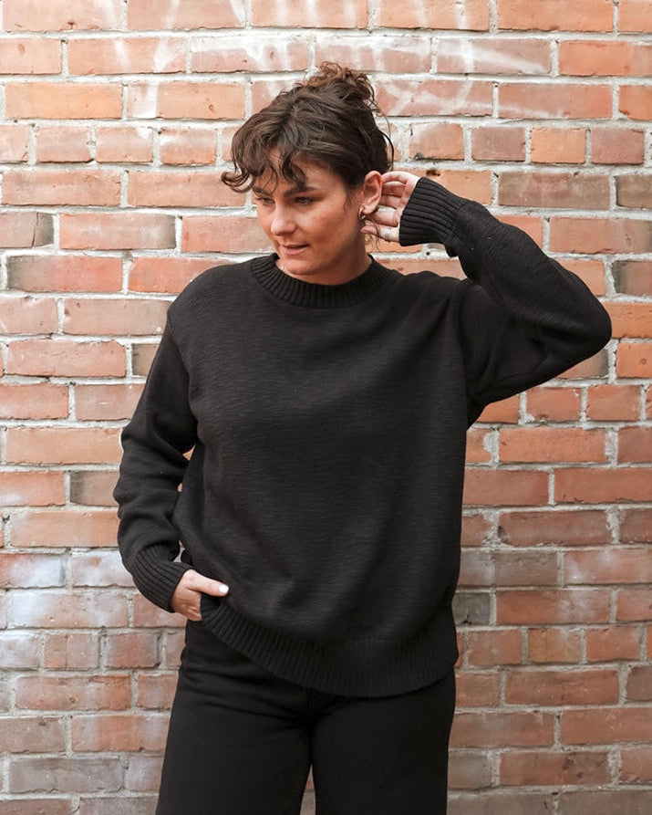A woman wearing a black knit crewneck sweater and black pants, posing against a red-brown brick wall.
