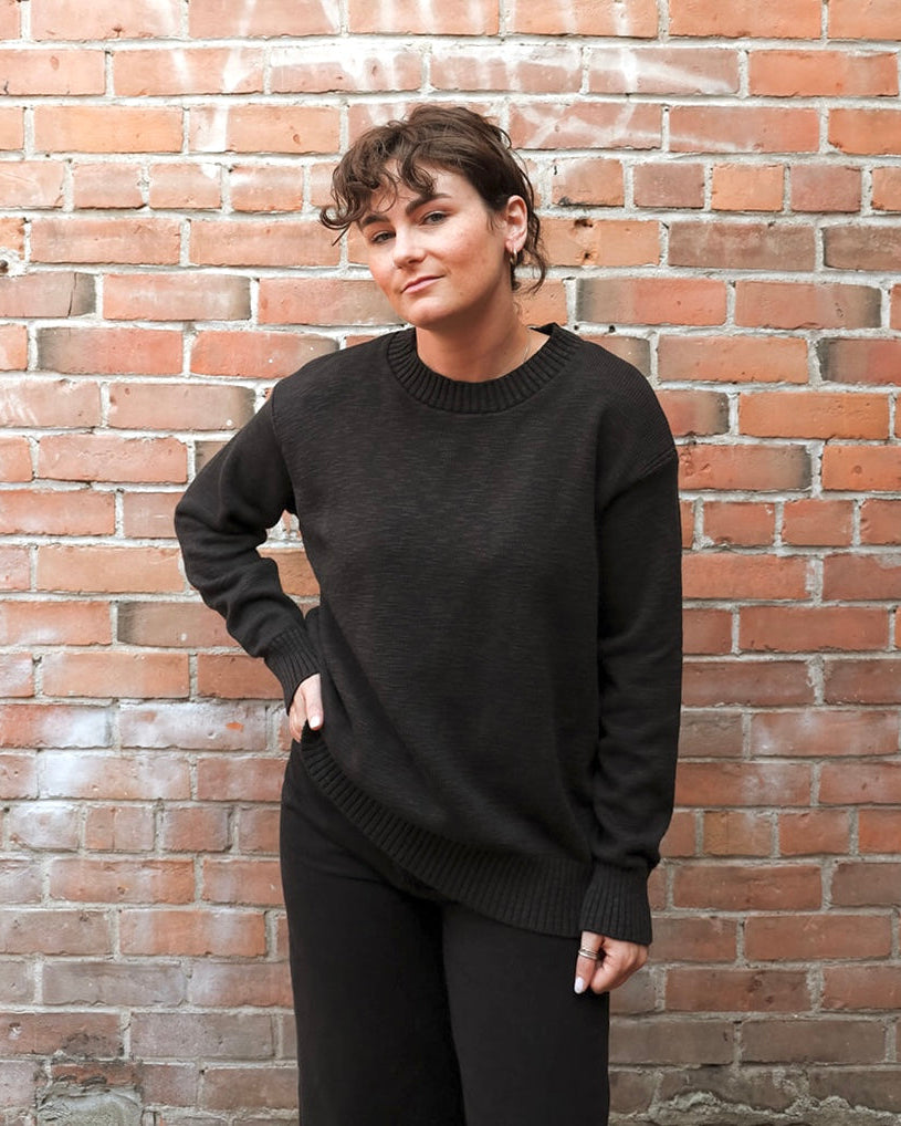 A woman wearing a dark charcoal knit crewneck sweater and black pants, standing against a red-brown brick wall.
