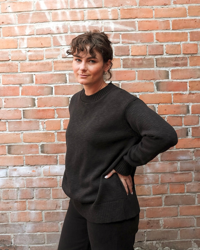 A woman wearing a dark charcoal knit crewneck sweater and black pants, posing with one hand in her pocket against a red brick wall background.