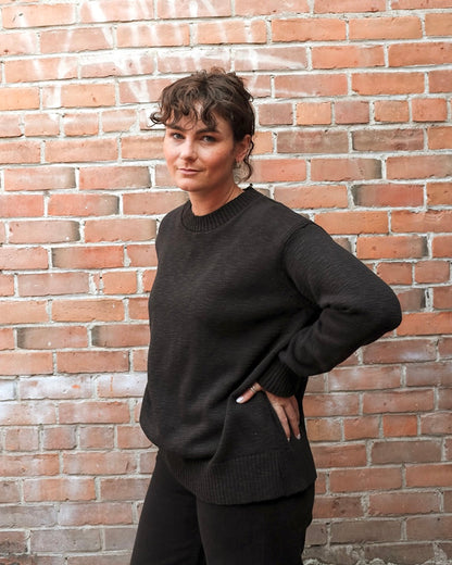 A woman wearing a dark charcoal knit crewneck sweater and black pants, posing with one hand in her pocket against a red brick wall background.