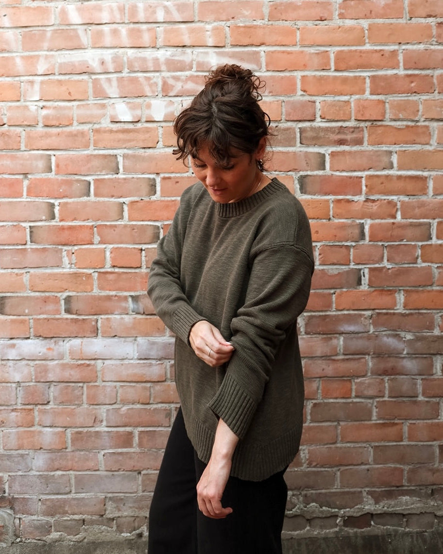A woman wearing an oversized olive green knit sweater and black pants, standing in front of a red brick wall.
