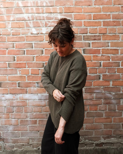 A woman wearing an oversized olive green knit sweater and black pants, standing in front of a red brick wall.