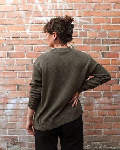 A woman viewed from behind wearing an olive green knit sweater and black pants, standing against a red brick wall.