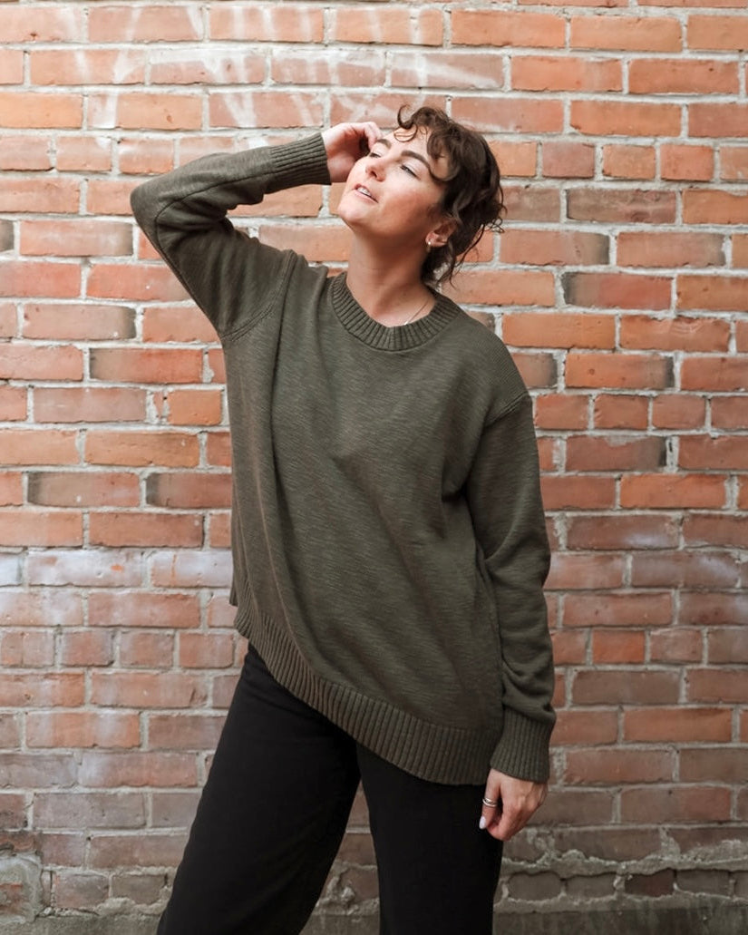 A person wearing an oversized olive green knit sweater and black pants, standing against a red-brown brick wall.