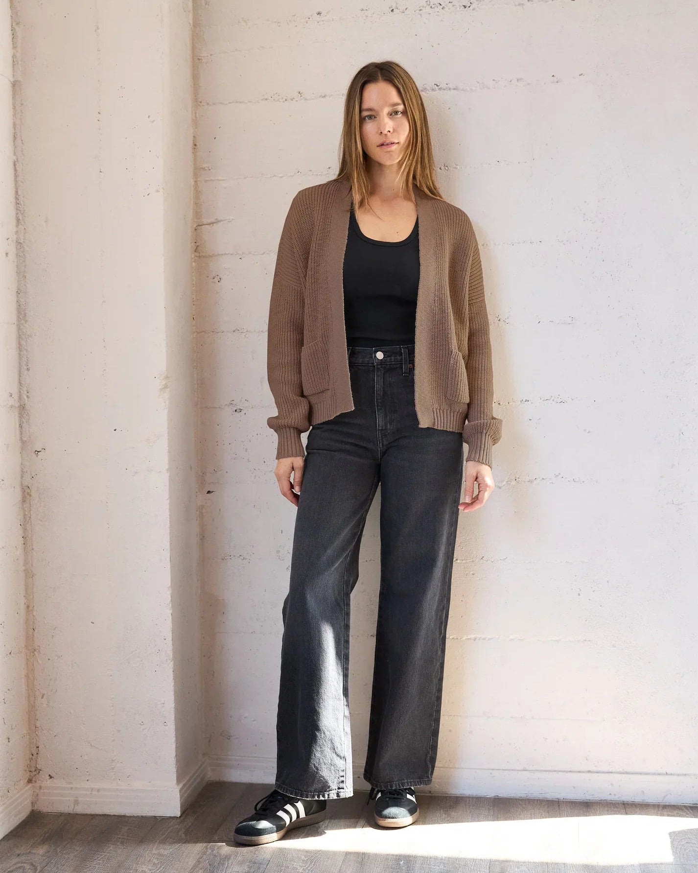 A woman stands in a bright, minimalist room with a white brick wall and wood floor, wearing a brown knit cardigan over a black tank top, black wide-leg jeans, and black sneakers.