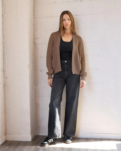 A woman stands in a bright, minimalist room with a white brick wall and wood floor, wearing a brown knit cardigan over a black tank top, black wide-leg jeans, and black sneakers.