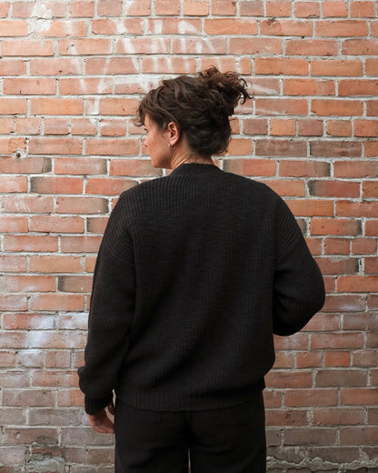 A person viewed from behind wearing a black textured knit sweater and black pants, standing in front of a red brick wall.