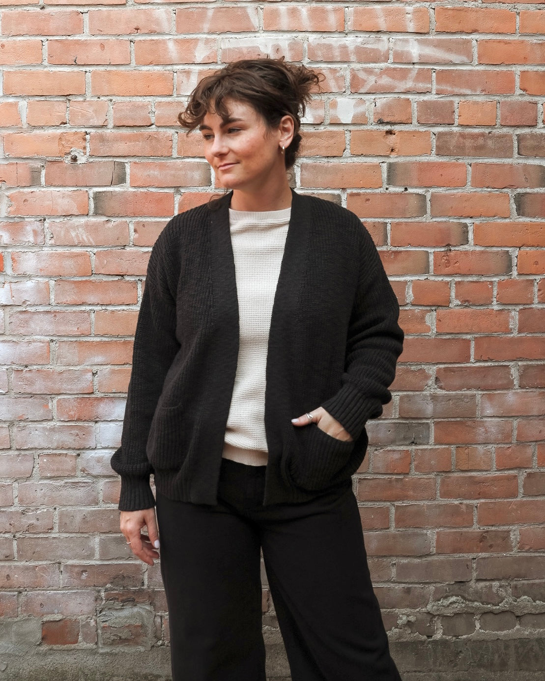 A woman wearing a black open-front knit cardigan with pockets, a cream ribbed top, and black wide-leg pants, standing against a red-brown brick wall.