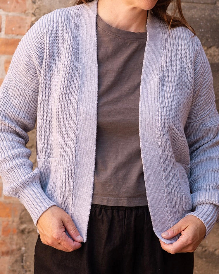 A woman wearing a light lavender ribbed knit cardigan over a brown t-shirt and black pants, standing in front of a rustic brick wall.