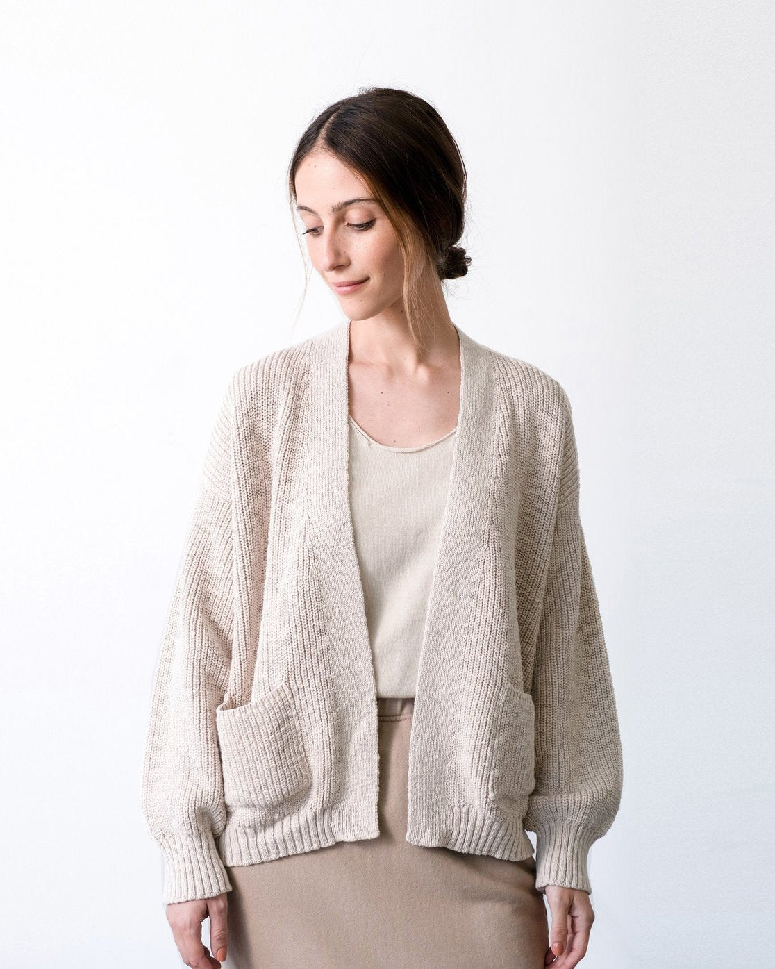 A woman wearing a light beige ribbed knit cardigan with front patch pockets over a matching beige top and taupe skirt, photographed against a clean white studio background.