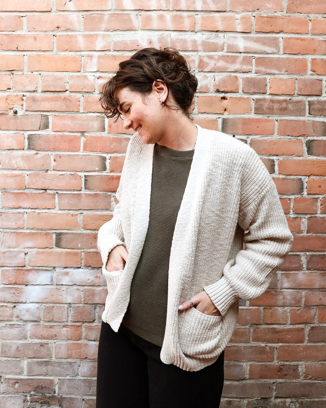 A woman wearing a cream-colored, open-front ribbed knit cardigan over a dark olive green top and black pants, standing against a red-brown brick wall.