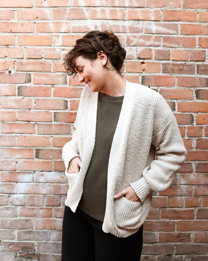 A woman wearing a cream-colored, open-front ribbed knit cardigan over a dark olive green top and black pants, standing against a red-brown brick wall.