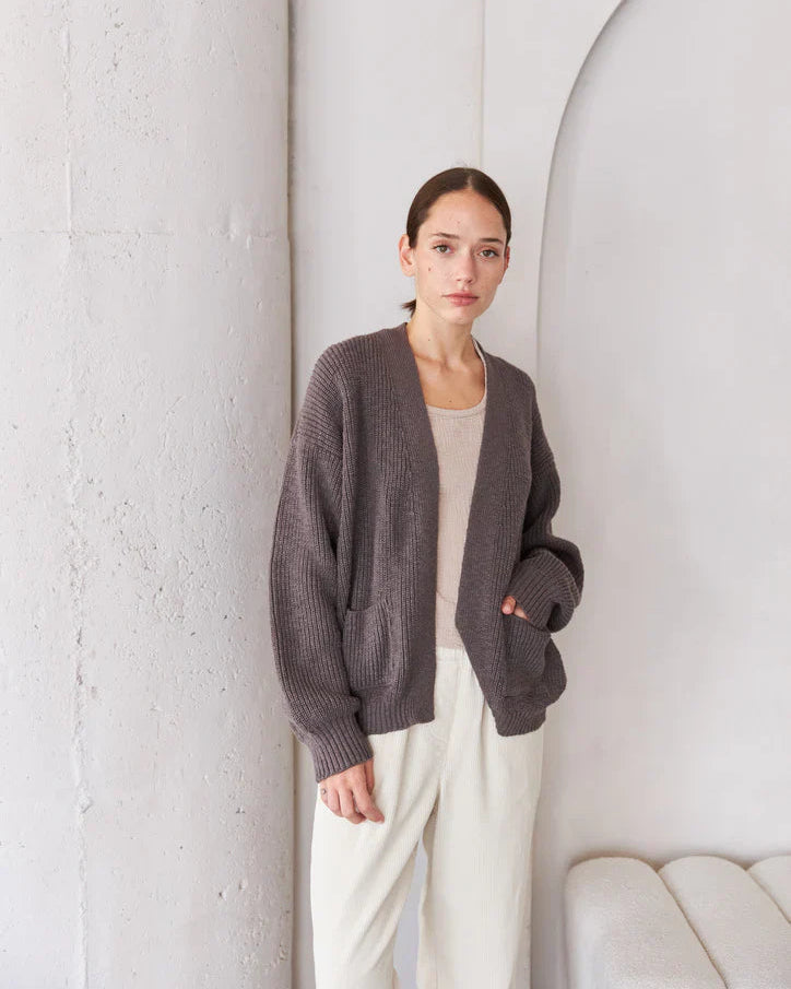 Woman wearing a dark gray cardigan over a white top and pants, standing against a white wall.