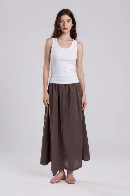 Woman wearing the It Is Well L.A Canada Everyday Skirt in Chocolate, a relaxed cotton maxi skirt with a drawstring waist, styled with a white ribbed tank and pink flats against a light grey studio background.
