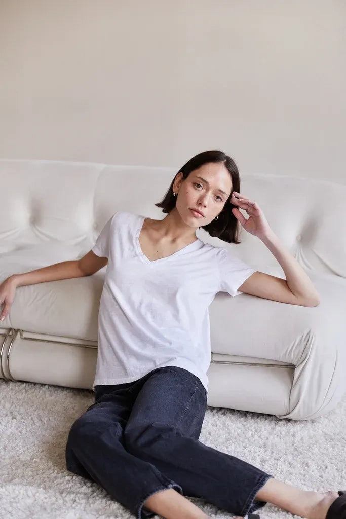 A woman with short dark hair sits on a white tufted sofa wearing a plain white V-neck t-shirt and dark blue jeans in a brightly lit, minimalist room.