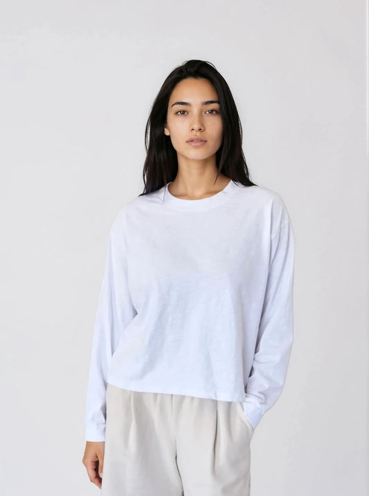 Woman wearing the It Is Well L.A Canada Long Sleeve Boxy Tee in Salt, a white organic cotton jersey top with a crew neck, dropped shoulders and a cropped straight hem, styled with light grey trousers against a pale grey studio background.