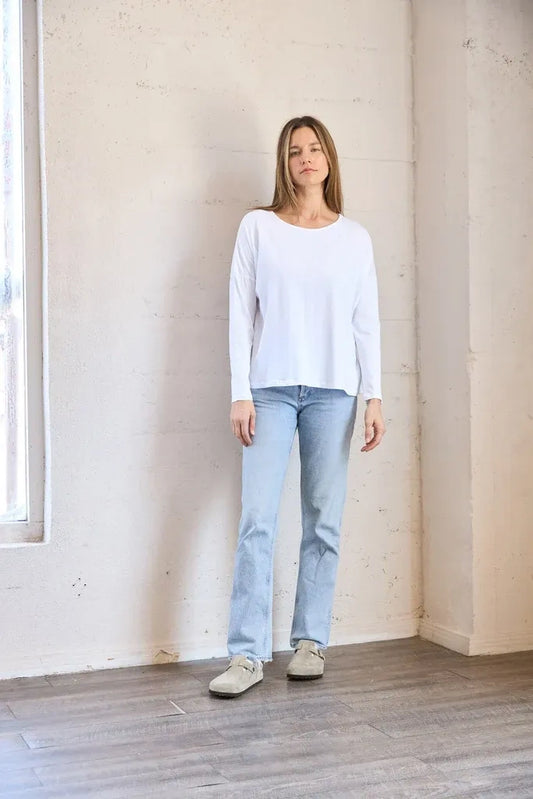 A woman wearing a white long-sleeve crewneck top, light blue jeans, and beige shoes, standing in a minimalist room with a concrete-textured wall and wood floor.