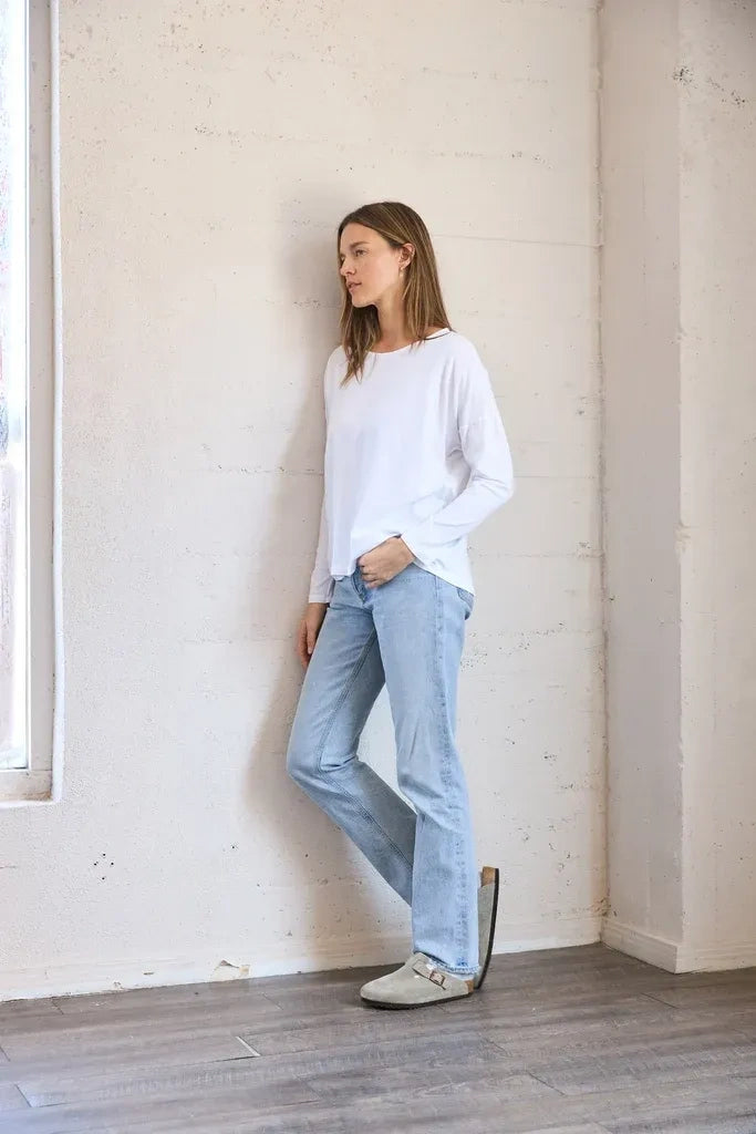 A woman stands in a minimalist room with a textured off-white wall and wood floor, wearing a white long-sleeve top, light-wash blue jeans, and beige loafers.