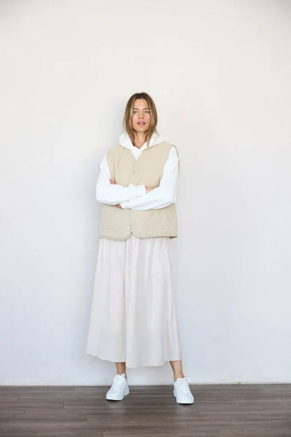 A woman stands with arms crossed wearing a beige quilted vest over a white long-sleeve top and a long white skirt, paired with white sneakers, against a plain off-white wall and dark wood floor.