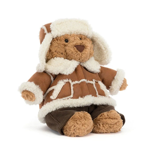 Jellycat Bartholomew Bear Alpine at an angle, a fluffy caramel bear in a cream-trimmed trapper hat, a tan shearling-look coat with cream piping and dark brown cord trousers, on a white background.