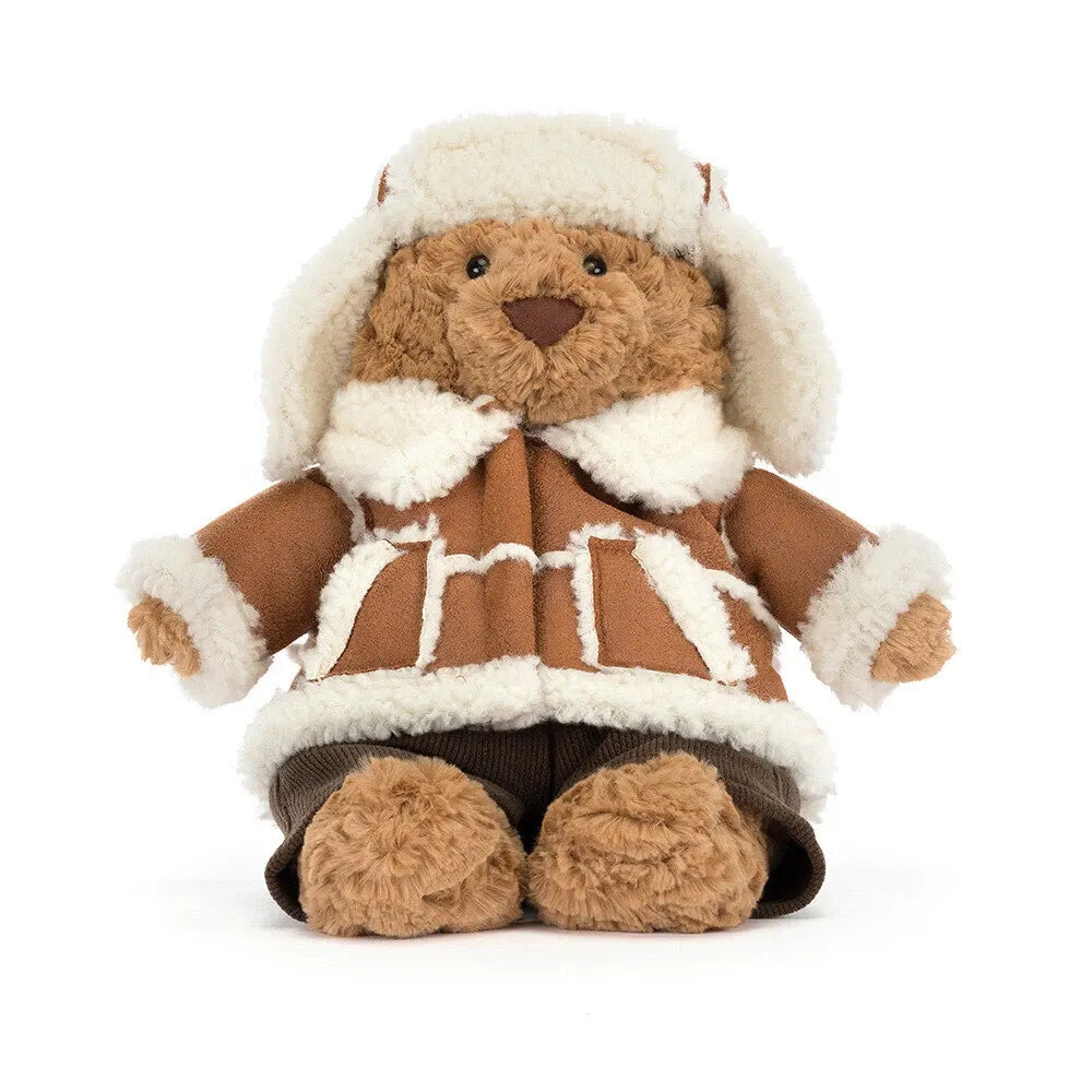 Front view of the Jellycat Bartholomew Bear Alpine sitting upright, dressed in a cream fleece-lined trapper hat, a tan coat with fluffy cream trim and pockets, and dark brown cord trousers.