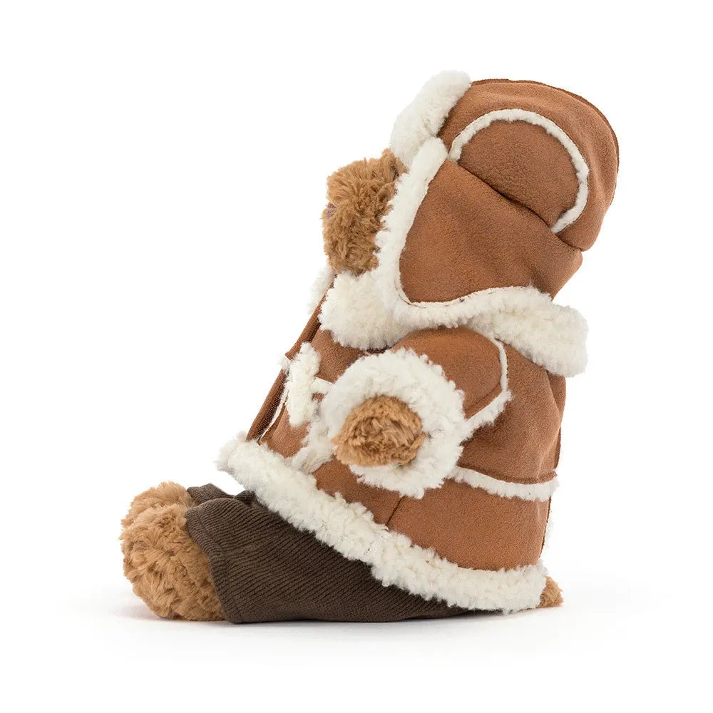 Side view of the Jellycat Bartholomew Bear Alpine, showing the trapper hat with its cream earflap, the tan coat with fluffy cuffs and the dark brown cord trousers.