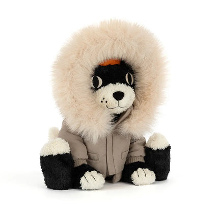Jellycat Jack Alpine at an angle, a black and cream plush cat with a freckled smile, wearing a taupe parka with a big fluffy cream faux-fur hood over an orange toque, on a white background.