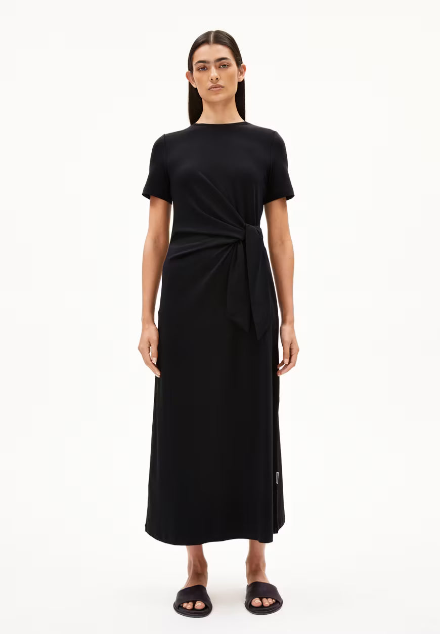 BLACK SHEATH WRAP DRESS ON MODEL WITH BLACK HAIR AND FLAT SANDALS AGAINST WHITE BACKGROUND