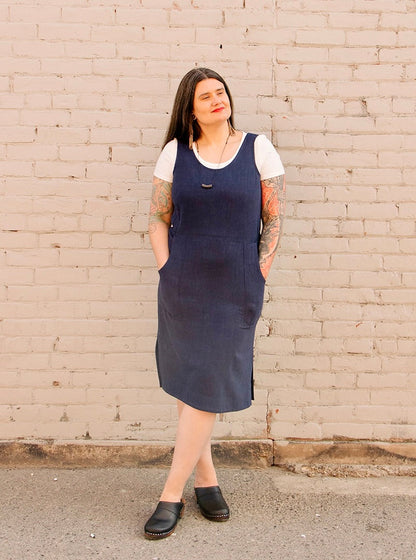 Woman wearing the Jennifer Glasgow Design Aisha Dress in Navy layered over a white tee, styled with a pendant necklace and black clogs, standing against a painted brick wall.