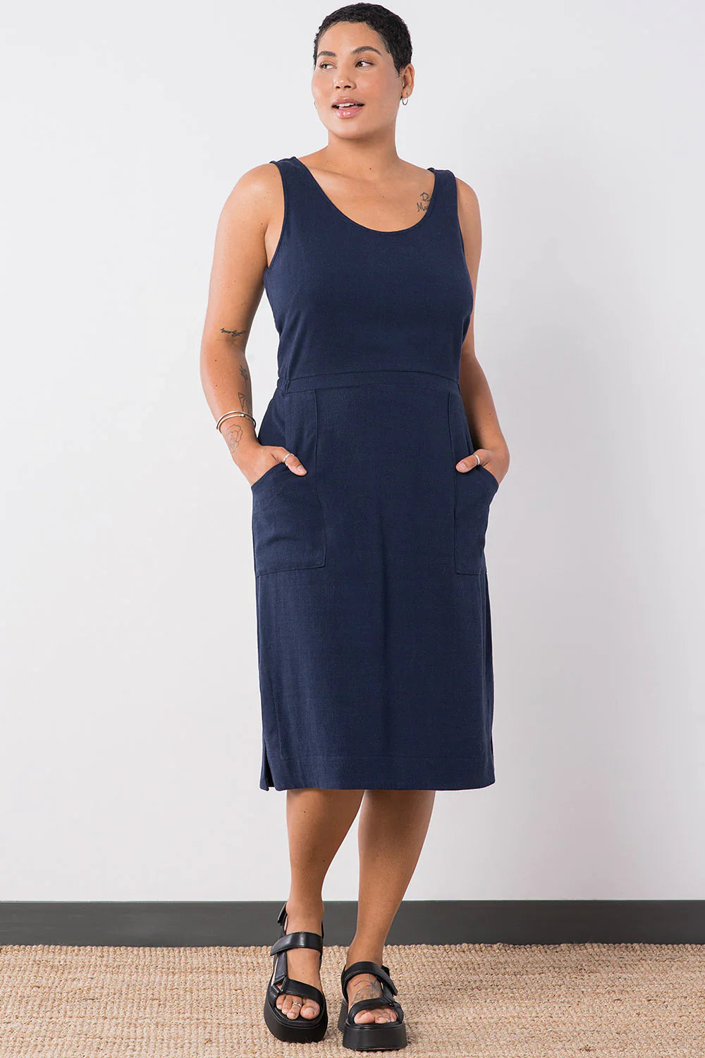 Woman wearing the Jennifer Glasgow Design Aisha Dress in Navy, a sleeveless scoop neck midi dress with a seamed waist and deep patch pockets, with both hands in the pockets and black sport sandals, against a white studio wall.